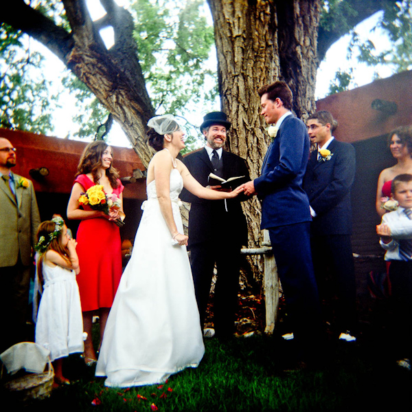 the happy couple exchanging vows during the ceremony under an oak tree - bride is wearing a white a-line dress with a birdcage veil and groom is wearing a blue suit with a yellow boutonniere - photo by New Mexico based wedding photographers Twin Lens